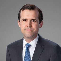 A man with short dark hair, exuding leadership, is wearing a navy blue blazer over a white dress shirt. He stands against a plain light gray background, facing the camera with a neutral expression.