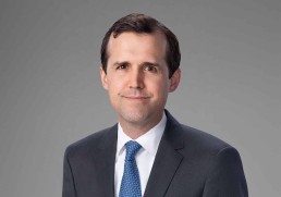 A man with short dark hair, exuding leadership, is wearing a navy blue blazer over a white dress shirt. He stands against a plain light gray background, facing the camera with a neutral expression.